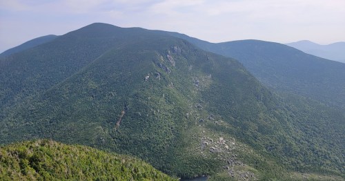 A panoramic view of lush green mountains under a clear sky, with rocky outcrops and a distant lake visible below.