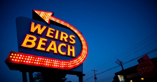 Neon sign for Weirs Beach with an arrow, illuminated against a twilight sky.