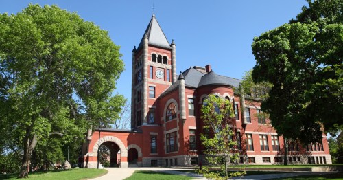 Historic red brick building with a clock tower, surrounded by green trees and a clear blue sky.