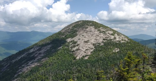 A rocky mountain peak surrounded by lush green forests under a partly cloudy sky.