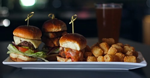 A platter featuring three sliders with toppings and a side of crispy tater tots, accompanied by a drink.