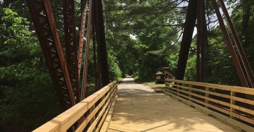 A wooden bridge with metal supports spans a path surrounded by lush green trees.