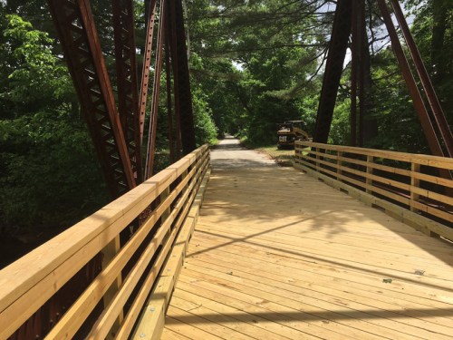 A wooden bridge with metal supports spans a path surrounded by lush green trees.