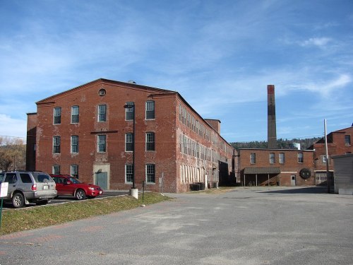 Historic brick factory building with a tall smokestack, set against a clear blue sky and surrounded by a parking area.