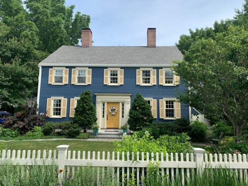 A blue two-story house with yellow shutters, a front door wreath, and a white picket fence surrounded by greenery.