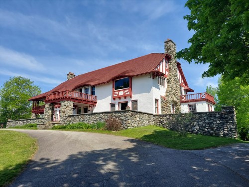A large, two-story house with a red roof and stone walls, surrounded by greenery and a clear blue sky.