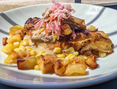 A plate of grilled fish topped with pickled vegetables, served with corn and crispy potatoes.
