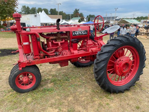A vintage red Farmall tractor displayed at an outdoor event, with people and tents in the background.