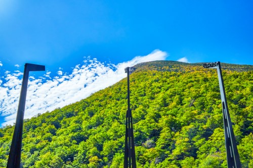 A vibrant green mountain under a bright blue sky with scattered clouds, framed by tall metal structures in the foreground.