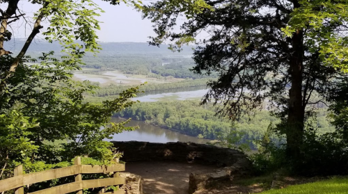 Scenic view of a river winding through lush greenery, framed by trees and a wooden railing in the foreground.