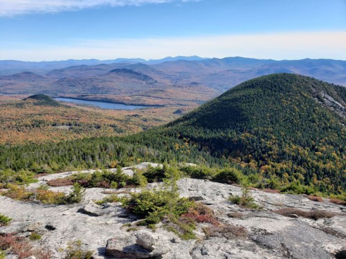 A panoramic view from a mountain summit, showcasing vibrant autumn foliage and a serene lake in the distance.