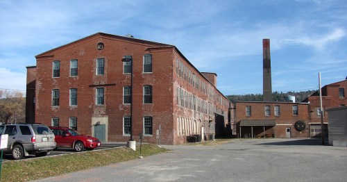 Historic red brick factory building with a tall chimney, surrounded by a gravel parking lot and blue sky.