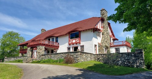 A large, charming house with a thatched roof, stone walls, and red accents, surrounded by greenery and a clear blue sky.