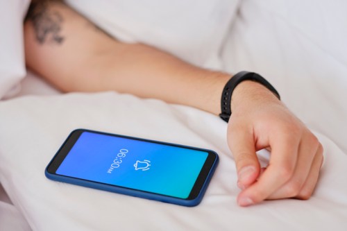 A hand rests on a bed next to a smartphone displaying an alarm time of 6:90.