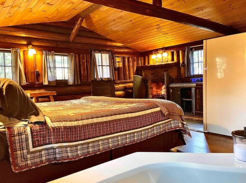 Cozy log cabin interior with a bed, fireplace, and rustic decor, featuring wooden beams and warm lighting.