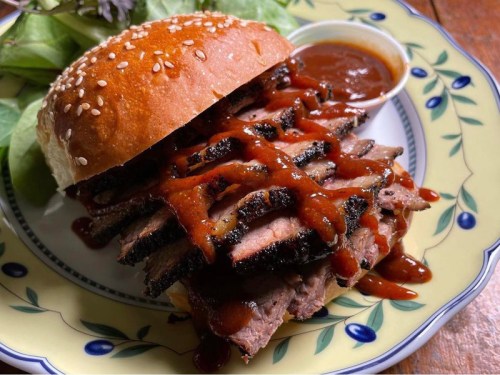 A close-up of a sesame seed bun filled with sliced brisket drizzled with barbecue sauce, served with a side of sauce.