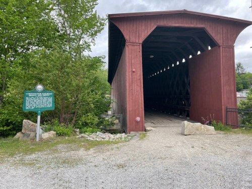 A red covered bridge entrance surrounded by greenery, with a historical sign nearby on a gravel path.