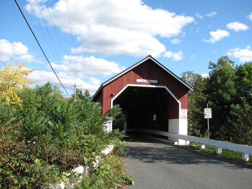 A red covered bridge surrounded by greenery and blue skies, with a road leading into the structure.