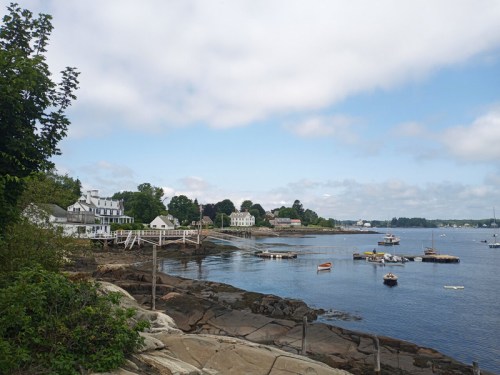 A serene coastal scene with boats on calm water, rocky shore, and charming buildings under a cloudy sky.