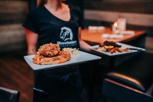 A server holds two plates of food: one with fried chicken and waffles, the other with assorted sides.