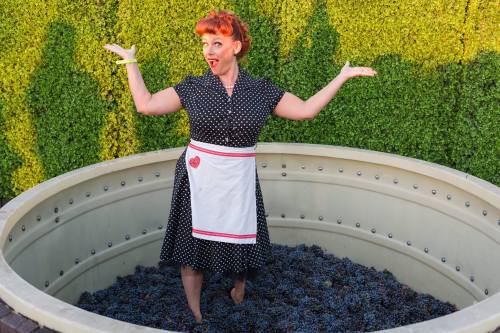 A woman in a polka dot dress stands in a large vat of grapes, smiling and gesturing playfully.