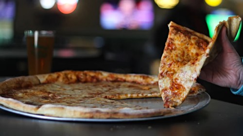 A hand lifts a slice of cheese pizza from a large pizza on a table, with a drink and blurred background.