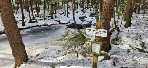 A snowy forest scene with a sign for William King Water Mill Site beside a partially frozen stream.