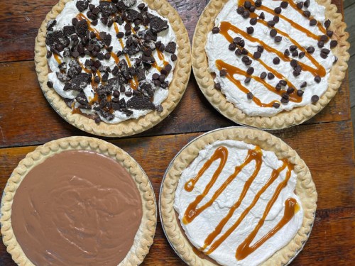 Four pies on a wooden surface: two with whipped cream and toppings, one chocolate, and one drizzled with caramel.