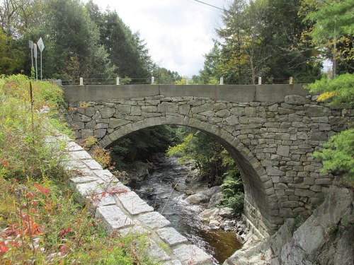 A stone arch bridge spans a small river, surrounded by trees and greenery under a cloudy sky.