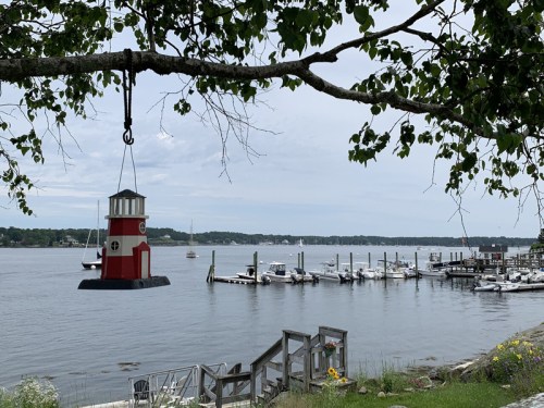 A red and white lighthouse hangs from a tree branch, overlooking a calm harbor with boats and a dock.