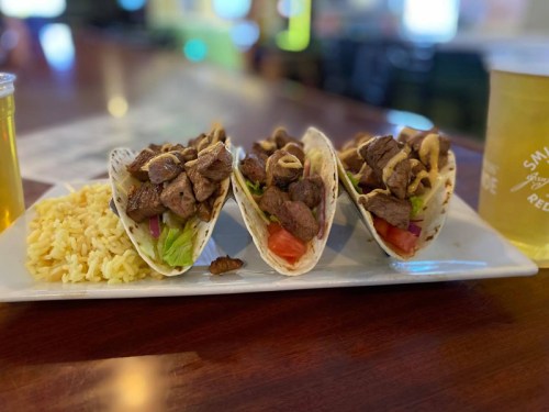 Three beef tacos filled with lettuce, tomato, and onions, served with a side of rice and two glasses of beer.