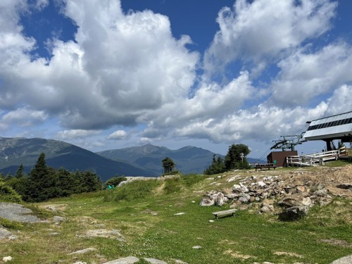 Scenic mountain landscape with green grass, rocky terrain, and a ski lift under a blue sky with fluffy clouds.