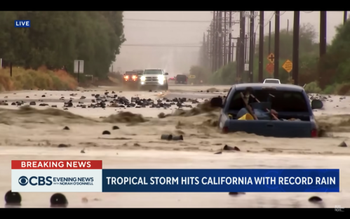 A flooded street in California with a submerged blue truck and heavy rain, as vehicles navigate through rising water.