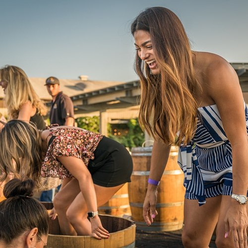 Two women are laughing and participating in a grape stomping event, surrounded by barrels and a festive outdoor setting.