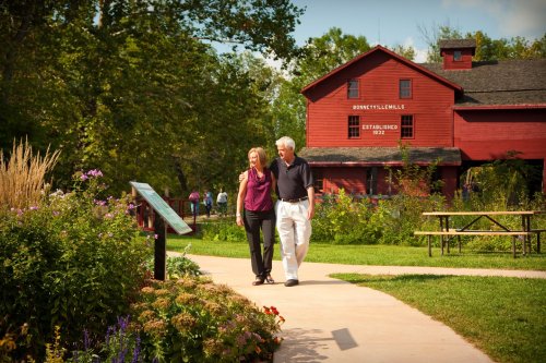 A couple walks hand in hand along a path in a park, with flowers and a red barn in the background.
