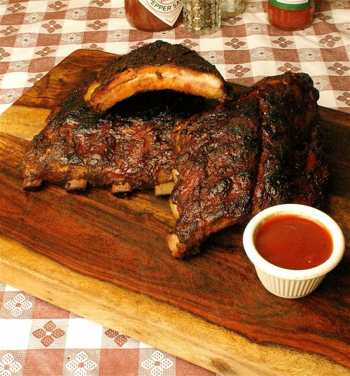 A wooden platter with grilled ribs and a small bowl of barbecue sauce, set on a checkered tablecloth.