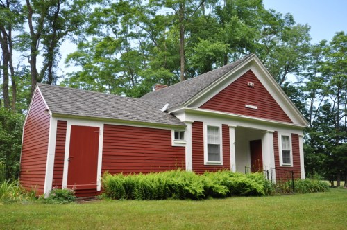 A charming red house with white trim, surrounded by greenery and trees, featuring a sloped roof and front porch.
