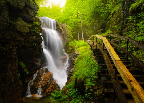 A serene waterfall cascades down rocky cliffs, surrounded by lush greenery and a wooden pathway.