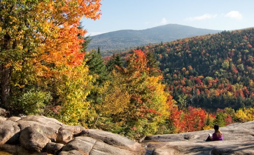 A person sits on rocks, surrounded by vibrant autumn foliage and mountains under a clear blue sky.