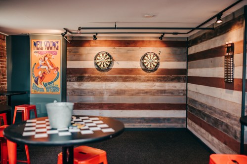 A cozy game room featuring dartboards on a wooden wall, a table with cards, and vibrant red stools.