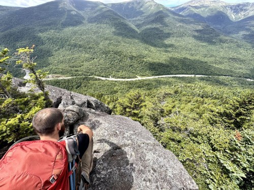 A hiker with a red backpack sits on a rocky ledge, overlooking a lush green valley and winding river below.