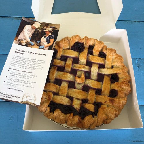 A freshly baked pie with a lattice crust sits in a white box next to a flyer about volunteering with Aurora Bakery.