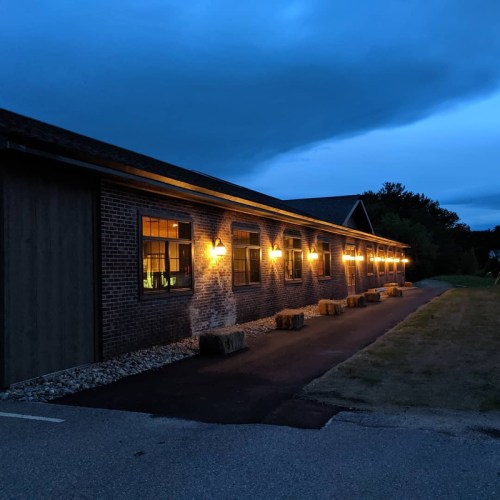 A brick building illuminated by warm lights at dusk, with hay bales lined along the pathway.