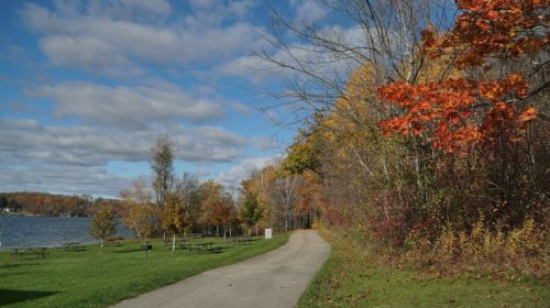 A scenic path along a lake, surrounded by colorful autumn trees and a partly cloudy sky.