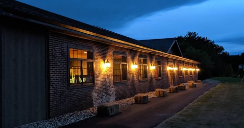 A brick building at dusk, illuminated by warm lights, with hay bales lined along the path.
