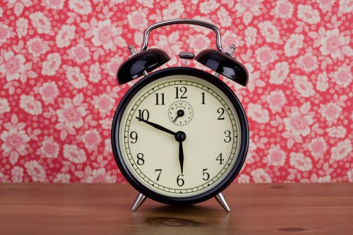 A vintage black alarm clock on a wooden table, set against a floral patterned pink background.