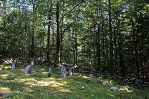 A serene forest scene featuring a small, old graveyard with several gravestones surrounded by trees and a stone wall.