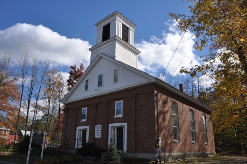 Historic brick building with a white bell tower, surrounded by autumn trees and a blue sky with clouds.