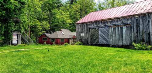 A red building and a weathered barn sit beside a lush green lawn, surrounded by trees under a clear sky.