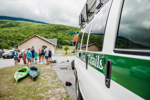 A group of people near kayaks and a vehicle with "Green Trails" branding, set against a scenic mountain backdrop.